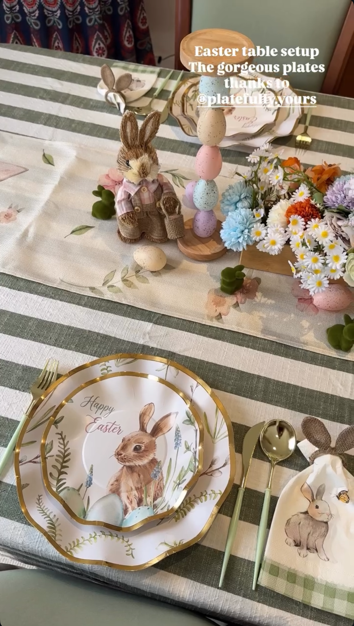 Easter table setting with decorative plates, bunny figurines, and flowers on a striped tablecloth.